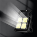 The black LED keychain light emits a strong white beam from its four yellow squares against a dark background.