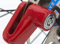 A close-up of a red motorcycle wheel security device clamped onto a bicycle disc brake, showing the silver lock mechanism.